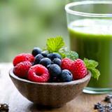Eine Schale mit frischen, bunten Beeren, Minze und Chia-Samen neben einem Glas grünen Smoothies auf einem Holztisch.